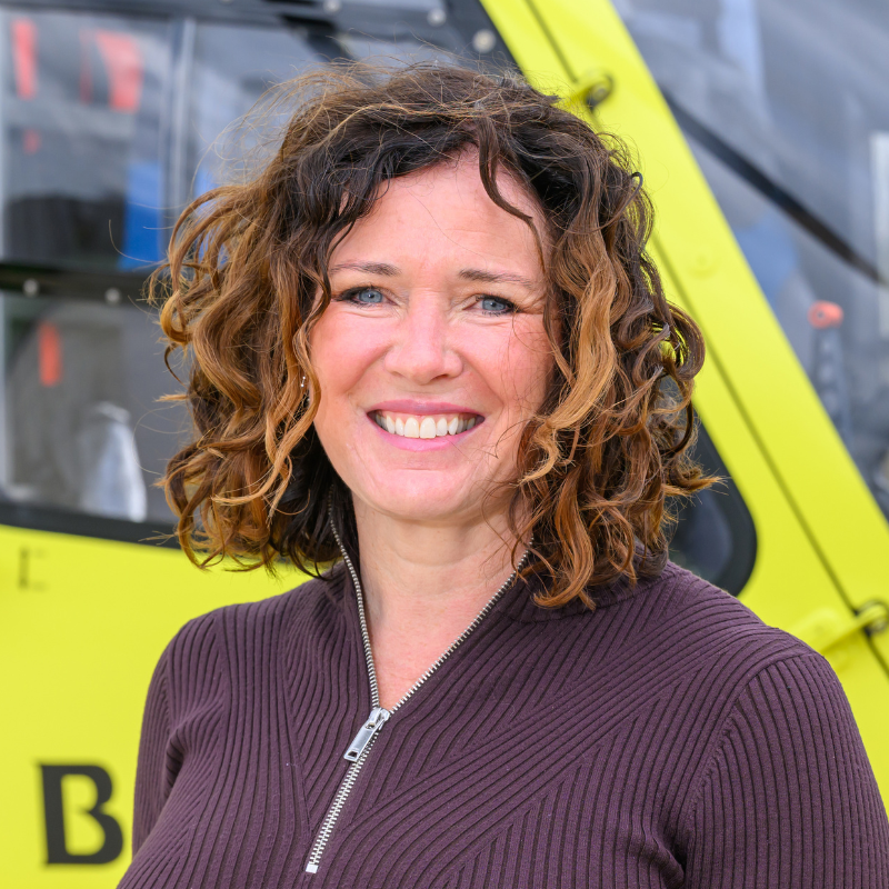Head and shoulders image of a women with shoulder length brown curly hair, wearing a brown top with silver zip, standing in front of a yellow helicopter.