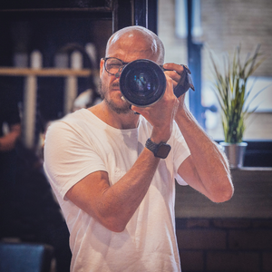 PBTH - Chris Whittam 300 x 300 px A man with short white hair, wearing a white short sleeved t-shirt is holding a camera up to his face as though taking a photo.