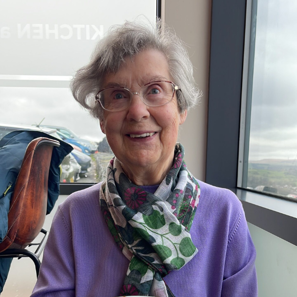 A lady with grey hair, wearing a lilac/purple cardigan and a patterned scarf.