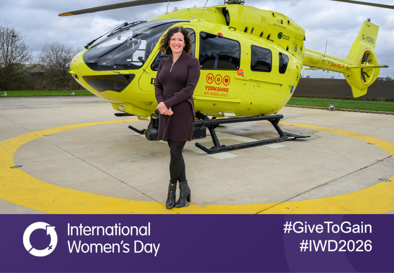 An image of women wearing a brown dress is stood in front of a yellow helicopter. There is a purple banner across the bottom with 'International Women's Day', '#IWD2026' and '#GiveToGain' in white