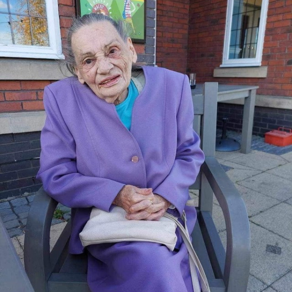 A lady with grey hair, wearing a lilac/purple suit and a light blue top, is sat in a dark grey garden chair. She has a light coloured handbag on her knee.