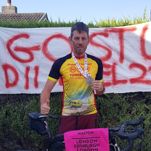 A man wearing a Yorkshire Air Ambulance cycling jersey and a medal, standing in front of a homemade banner.