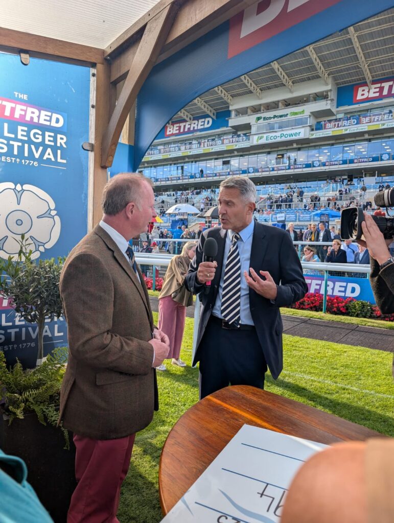 An interview between two men stood on or next to a racecourse