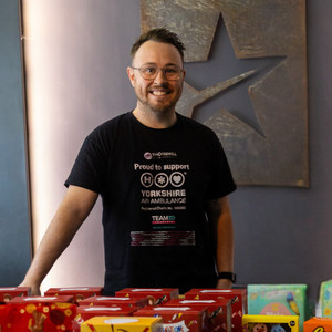 A man with short dark hair, wearing glasses and a black t-shirt is stood in front of a table full of easter eggs.