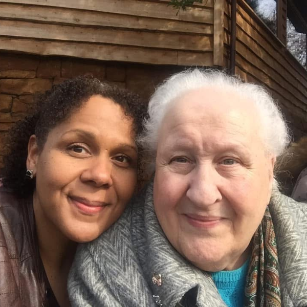An image of two ladies. One with white hair, wearing a patterned grey coat with two brooches on the collar, a patterned scarf and a blue jumper. The other lady has dark hair, and is wearing a brown leather jacket . She is stood close to the first lady and has her head gently on her shoulder.