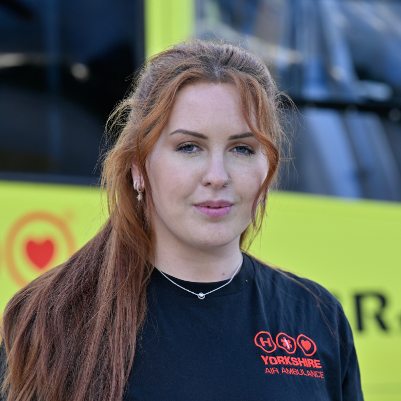 A person with long red hair, wearing black top with red logo is standing in front of a yellow helicopter