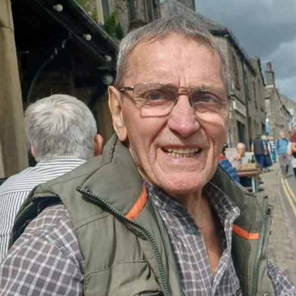 A man with short grey hair, wearing glasses, a checked shirt and a green gilet.