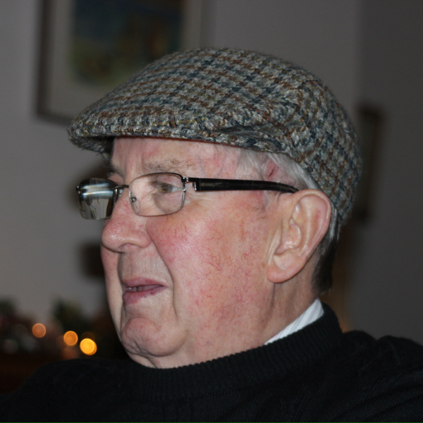 A side view of a man with grey hair, wearing glasses and a flatcap.