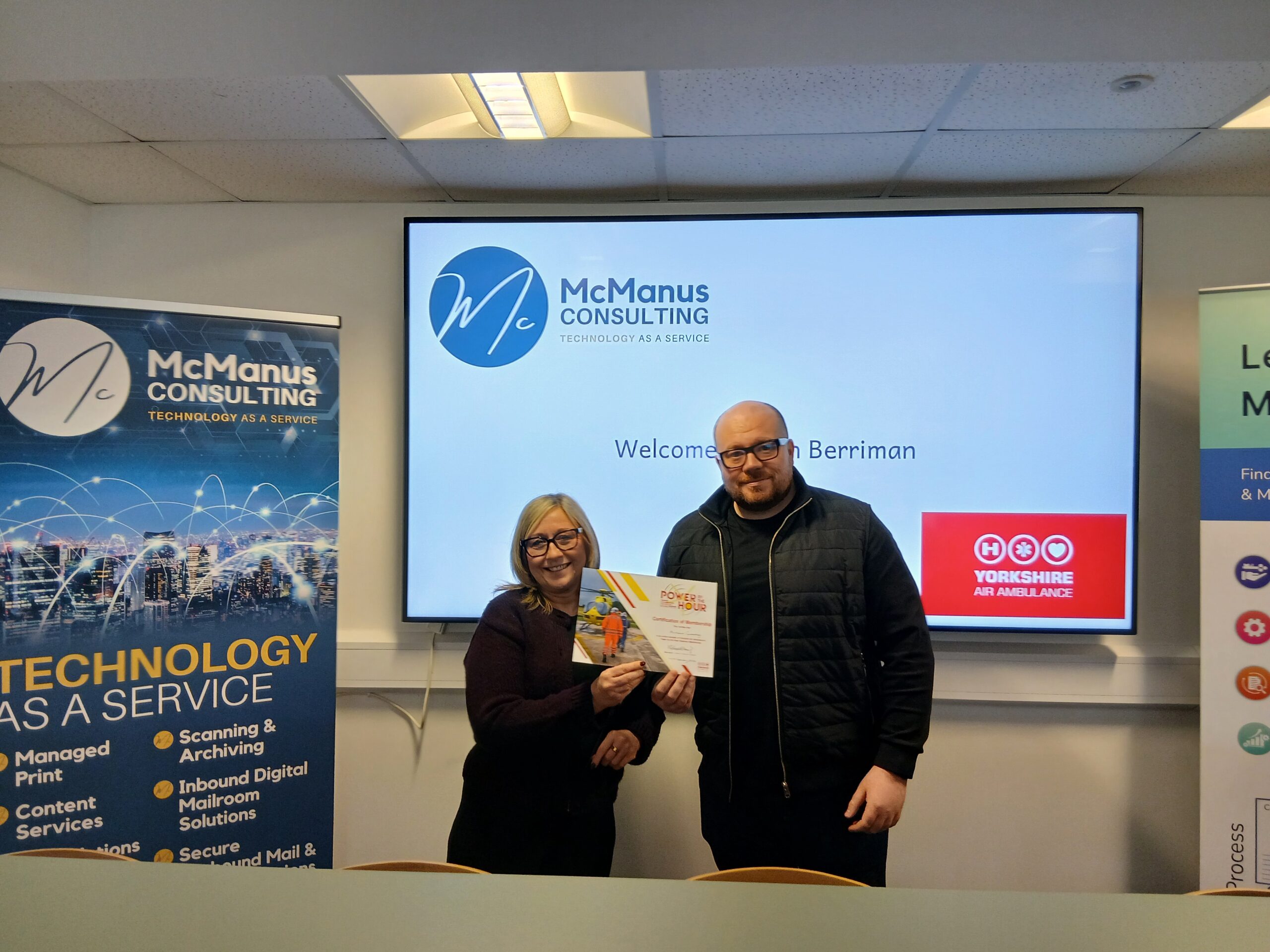 McManus Consulting A woman with blonde hair and glasses and a man with glasses are stood in front of a digital presentation board holding a certificate.