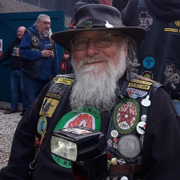 A man with a long white beard and white moustache, wearing black leather waistcoat which ahs lots of badges sewn on to it. He has a black top under the waistcoat and is wearing a black had with pin badges on it.