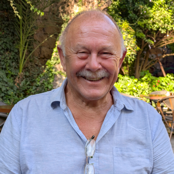 A man with a grey moustache, wearing a light blue linen shirt with a pair of glasses hanging on the shirt.