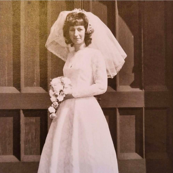 A black and white photo of a lady in a white wedding dress with a white veil. She is holding a bouquet of flowers and is stood in front of a panelled wall.