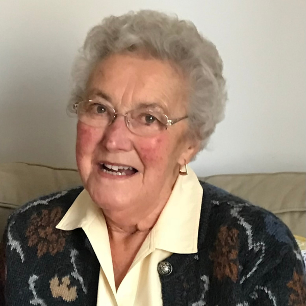 A lady with curly grey hair, wearing glasses, gold earrings, a light yellow blouse and a dark patterned cardigan.