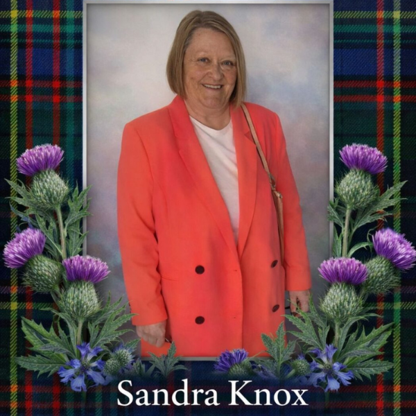 A lady with bobbed light brown/blonde hair, wearing a white top and orange/coral jacket. The image has a border which has a tartan pattern and thistles. At the bottom is the name 'Sandra Knox'