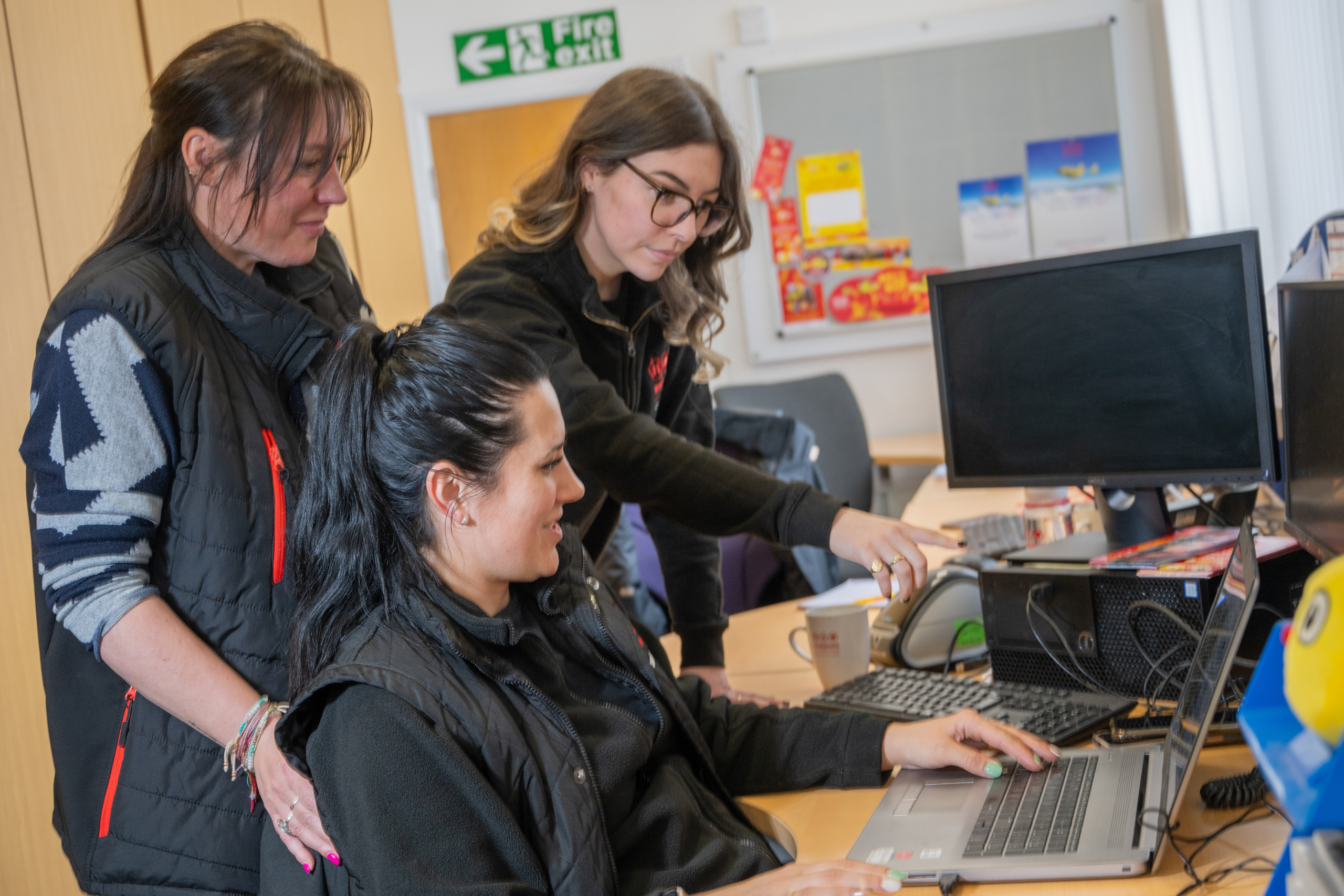 A lady is sat at a desk with a laptop and screen and two ladies are stood behind her.