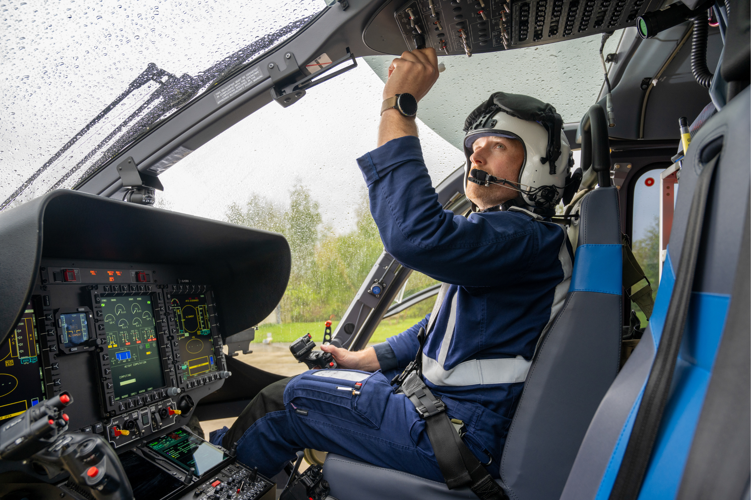 A man wearing a dark blue flight suit and white helmet sat in a helicopter cockpit adjusting some of the overhead instruments