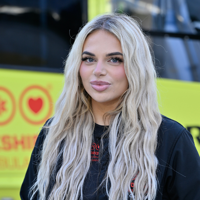 A person with long blonde hair, wearing black top with red logo is standing in front of a yellow helicopter