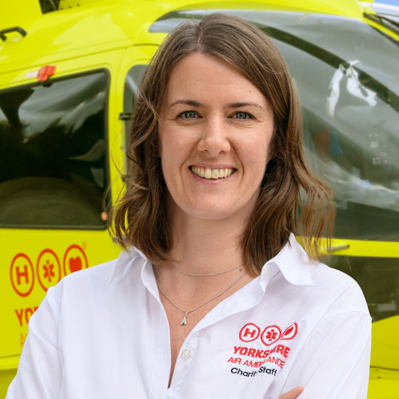 A lady with shoulder length brown hair, wearing a white shirt with a red logo, is standing in front of a yellow helicopter.
