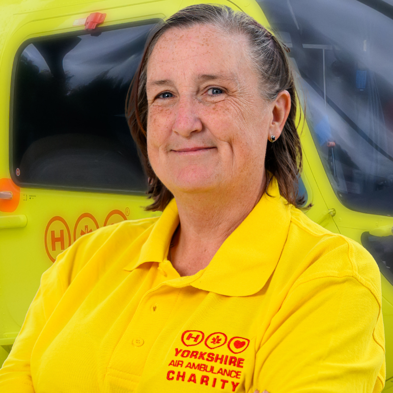 A lady wearing a yellow t-shirt featuring a red logo is stood in front of a yellow helicopter.
