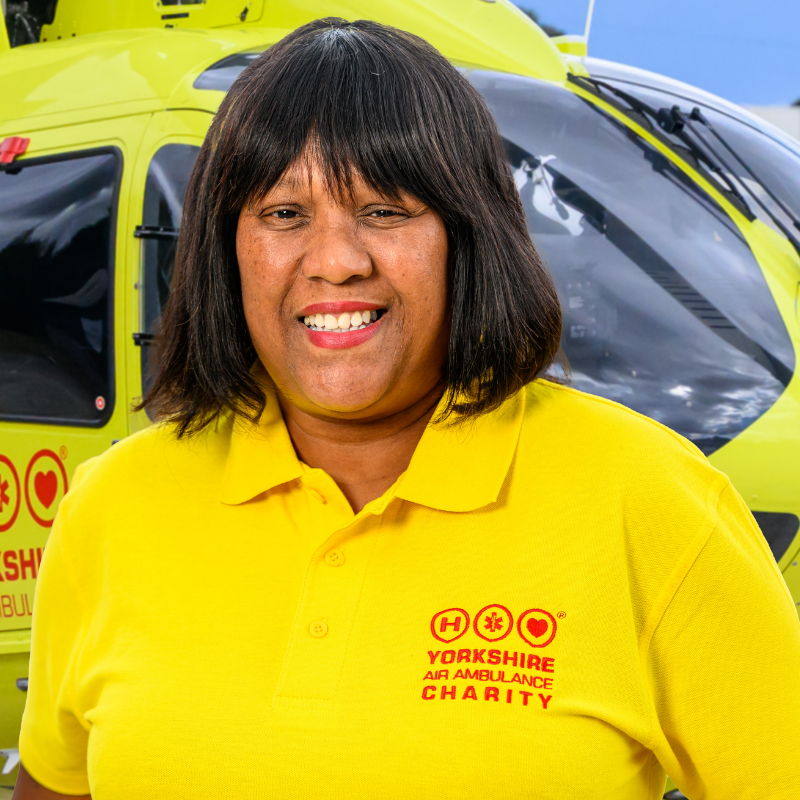 A lady wearing a yellow t-shirt featuring a red logo is stood in front of a yellow helicopter.