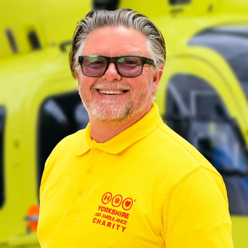 A man wearing glasses and a yellow t-shirt featuring a red logo is stood in front of a yellow helicopter.