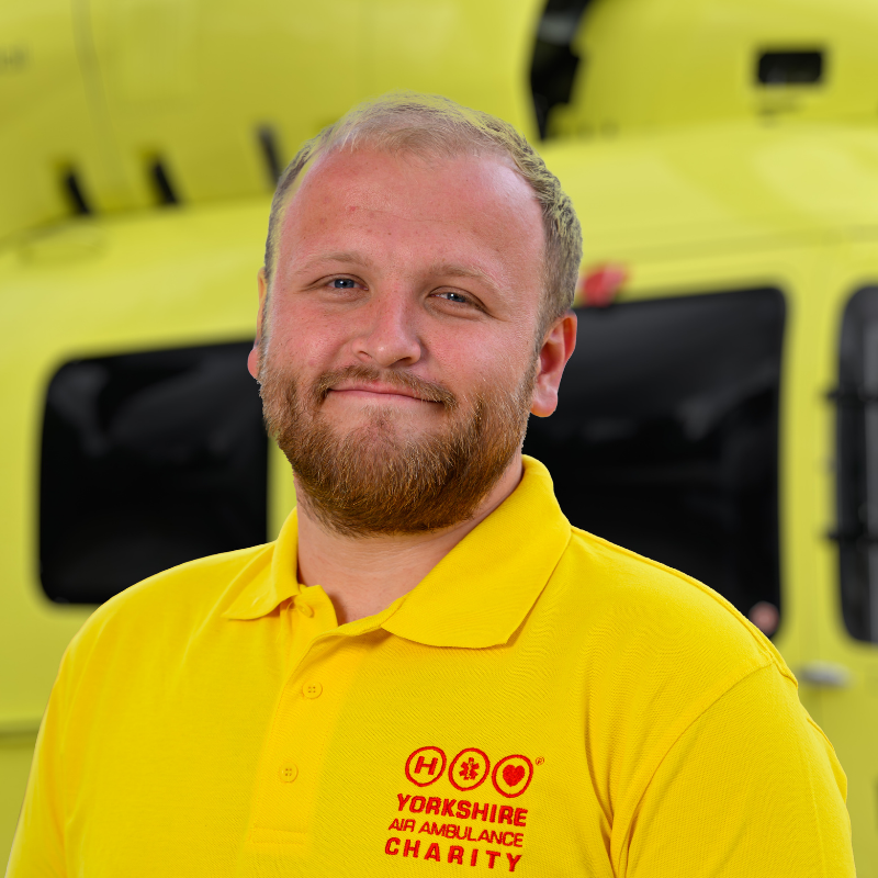 A man wearing a yellow t-shirt featuring a red logo is stood in front of a yellow helicopter.