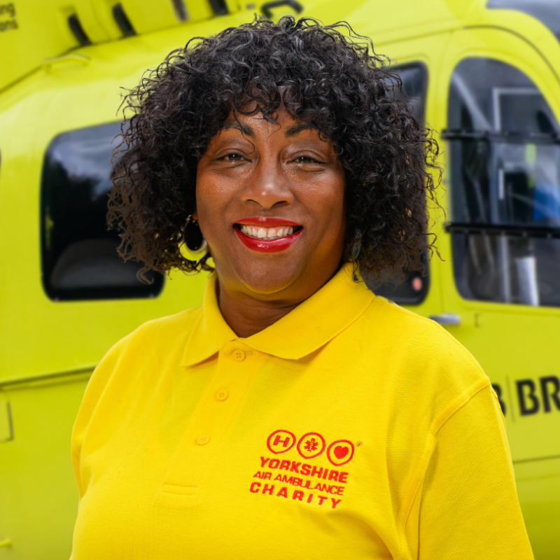 A lady wearing a yellow t-shirt featuring a red logo is stood in front of a yellow helicopter.