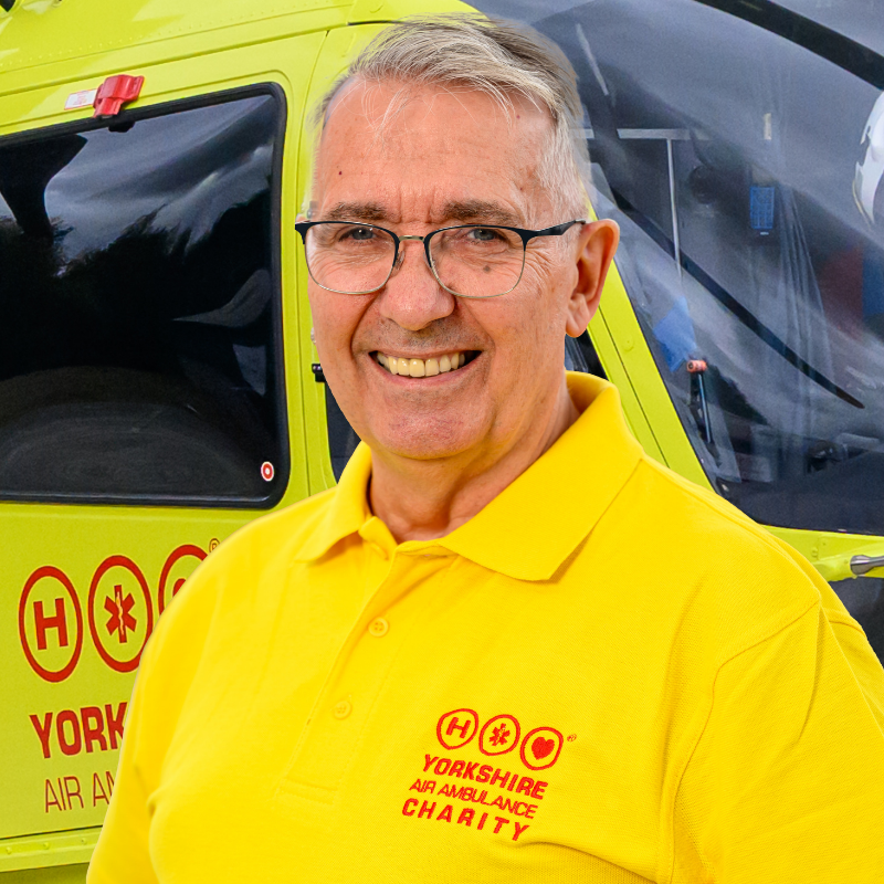 A man wearing glasses and a yellow t-shirt featuring a red logo is stood in front of a yellow helicopter.