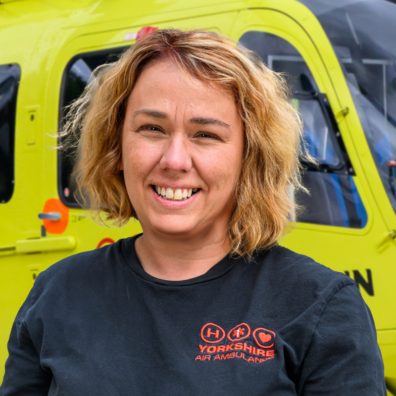 A lady with shoulder length blonde hair, wearing a black top with a red logo, is standing in front of a yellow helicopter.