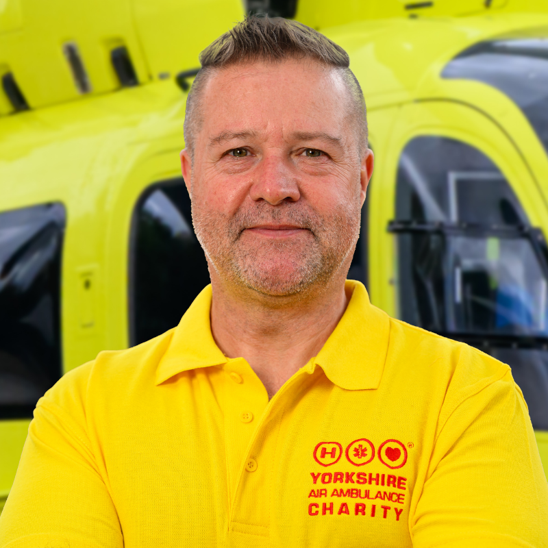 A man wearing a yellow t-shirt featuring a red logo is stood in front of a yellow helicopter.