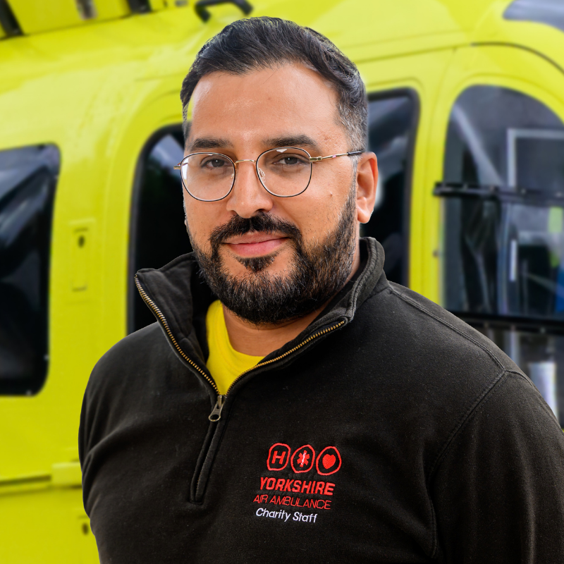 A man with short dark brown or block hair, wearing a black zip up top with a red logo, is standing in front of a yellow helicopter.
