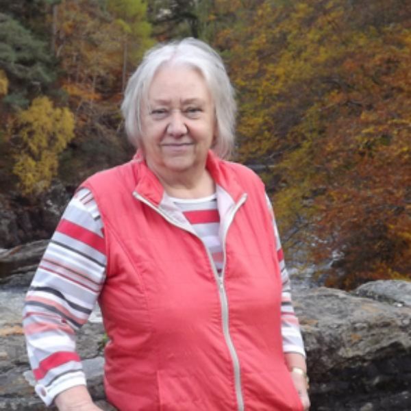 A lady with shoulder length hair, wearing a pink gilet and striped long sleeved top.
