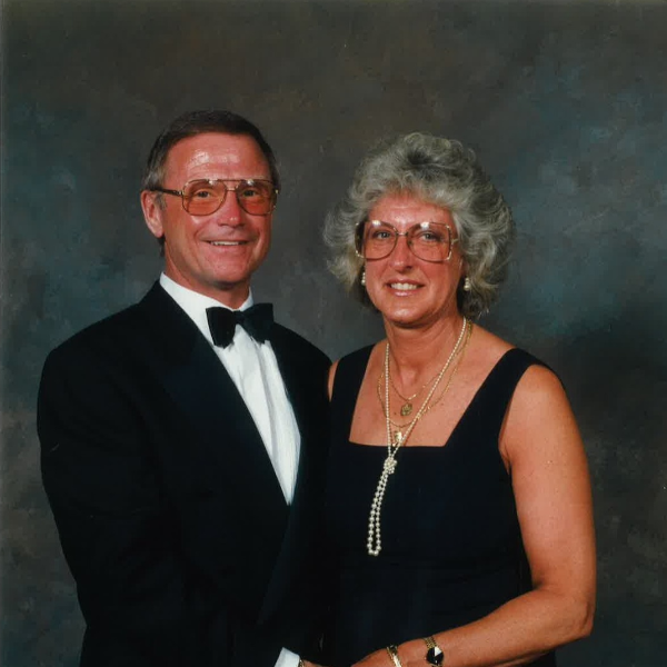 A man with glasses and a black suit, with white shirt and black bow tie is stood with a lady with glasses wearing a black dress. A professional photo which looks to have been taken at a dinner event.
