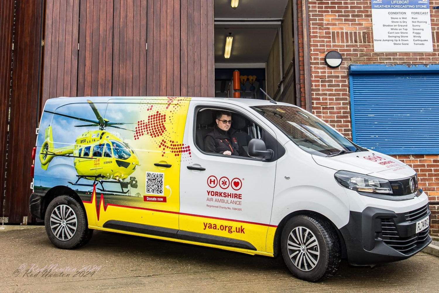 YAA Website Block Image - Fundraising Van - 1500 x 500 px A man wearing glasses and a black top is sat in a white van which has graphics featuring a yellow helicopter.
